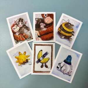 May include: Six rectangular art prints featuring watercolor illustrations: a monkey hugging a plush toy, an orangutan holding a baby, a bee, a smiling sun, a banana taped to a frame, and a pigeon. Each print has a white border and soft colors.