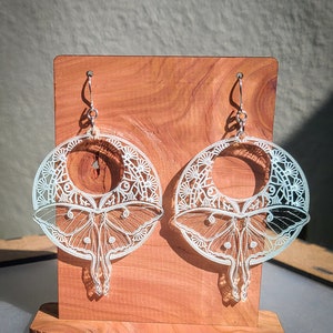 May include: A pair of silver earrings with a moth design. The earrings are laser-cut with intricate details, including a circular frame with geometric patterns and a moth silhouette. The earrings are displayed on a wooden stand.