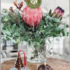 Puede incluir: Dos adornos de madera con forma de árbol de Navidad y bola, ambos hechos de cápsulas de semillas de Banksia. Los adornos están sobre una mesa de madera con un ramo de flores rosas y moradas en un jarrón de vidrio.