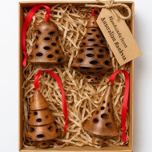 Op de afbeelding: Een set van vier bruine Banksia-ornamenten in een kartonnen doos met rode linten. De ornamenten hebben een bel- en boomvorm met gaten. Het label luidt "Handmade from Australian Banksia".