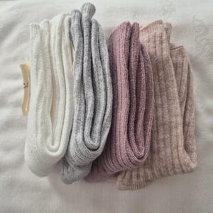 May include: Four pairs of folded ribbed socks in a variety of neutral colors: white, gray, mauve, and beige. The socks are neatly arranged on a white surface, showcasing their texture and color variations. A small tag is visible.