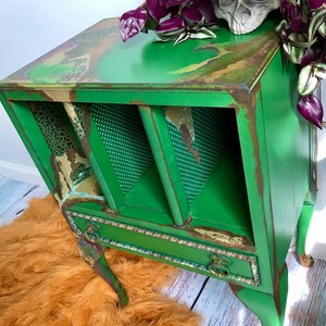 May include: A green painted side table with a distressed finish. The table has a drawer and two open shelves. The table is sitting on a fluffy orange rug.