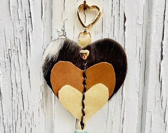 May include: A layered heart-shaped keychain made of brown, tan, and white cowhide leather with a gold metal clasp and a turquoise bead on a brown and tan leather tassel.