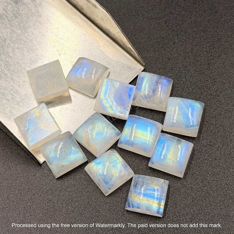 May include: A collection of twelve square-cut moonstone cabochons with a translucent white appearance and iridescent blue flashes, displayed on a metallic scoop. Each gemstone features a smooth, polished surface and a flat base.