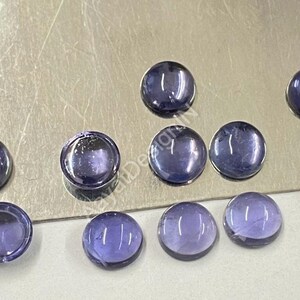 May include: Ten round, smooth, purple gemstones with a slight shimmer. The gemstones are arranged in a semi-circle on a silver surface.
