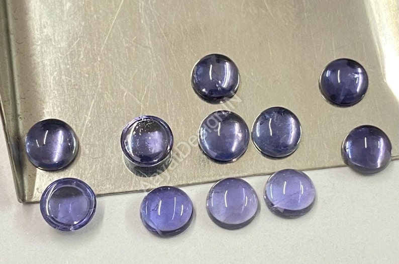 May include: Ten round, smooth, purple gemstones with a slight shimmer. The gemstones are arranged in a semi-circle on a silver surface.