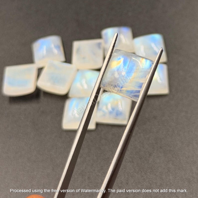 May include: A collection of square, translucent moonstone cabochons with iridescent blue and white flashes. One square stone is held by metal tweezers, displaying a smooth surface and internal light reflections, while others are scattered behind.