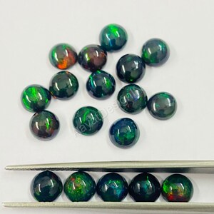 May include: A collection of round, black opal cabochons with green and red flashes of colour. The cabochons are scattered on a white surface and some are held in tweezers.