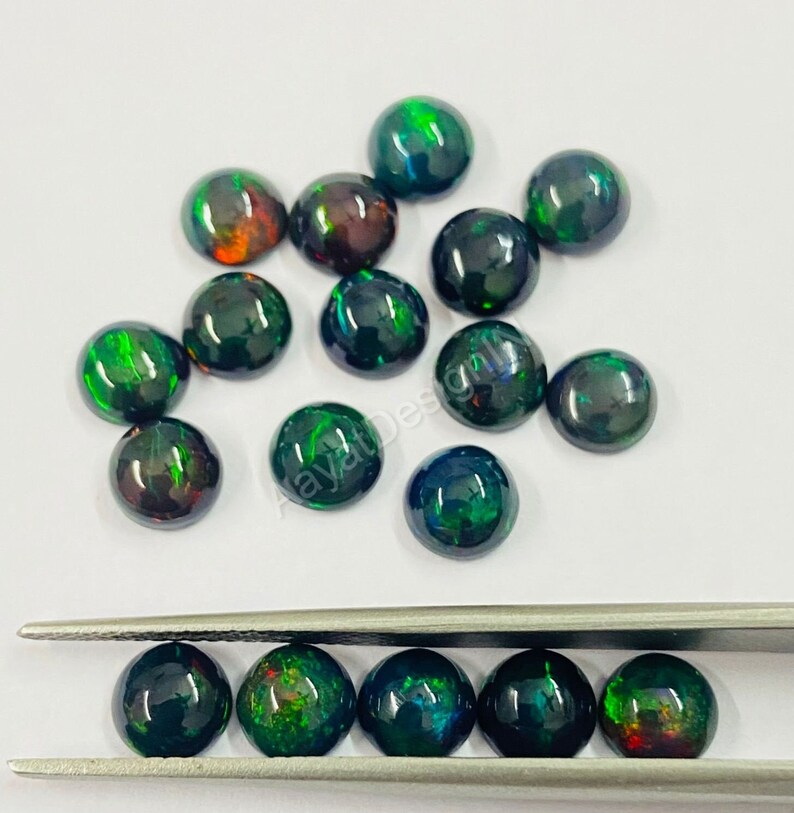 May include: A collection of round, black opal cabochons with green and red flashes of colour. The cabochons are scattered on a white surface and some are held in tweezers.