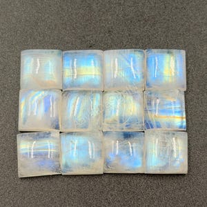 May include: A grid of twelve square moonstone cabochons arranged in three rows of four. Each translucent stone displays a distinct blue adularescence and internal light reflections, with a smooth, polished surface and rounded edges.