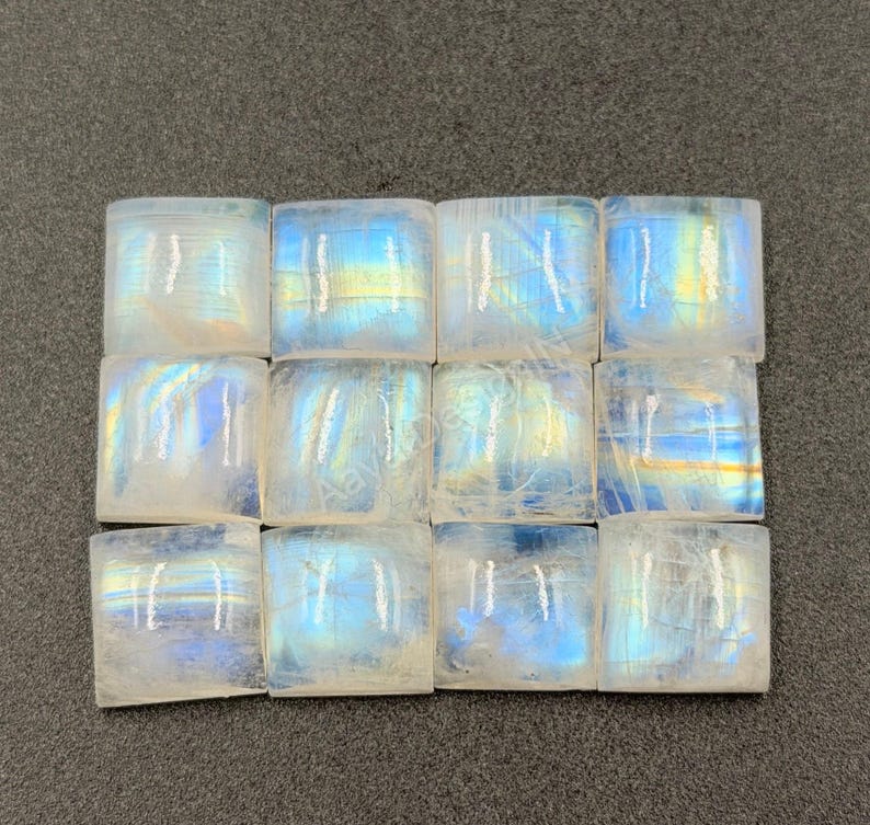 May include: A grid of twelve square moonstone cabochons arranged in three rows of four. Each translucent stone displays a distinct blue adularescence and internal light reflections, with a smooth, polished surface and rounded edges.