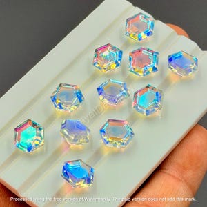 May include: Iridescent, hexagonal-shaped crystals arranged on a white surface. The crystals reflect a spectrum of colors, including blue, pink, and yellow. The crystals are likely decorative elements for crafts or jewelry making.