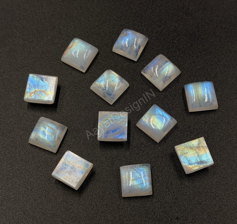 May include: Eleven square-shaped moonstone cabochons with a smooth, polished surface. Each stone displays a translucent white base with iridescent blue and yellow flashes of light. The stones are arranged in a scattered pattern.