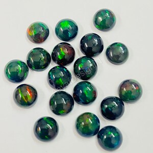 May include: A collection of 16 black opal cabochons with green and red iridescent flashes. The cabochons are round and have a smooth, polished surface.