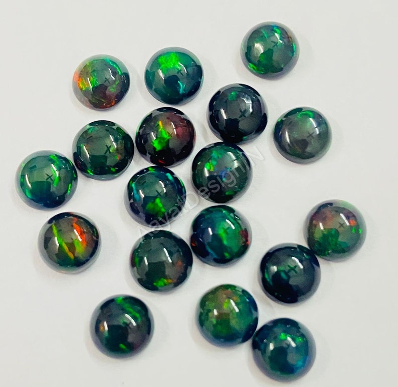 May include: A collection of 16 black opal cabochons with green and red iridescent flashes. The cabochons are round and have a smooth, polished surface.