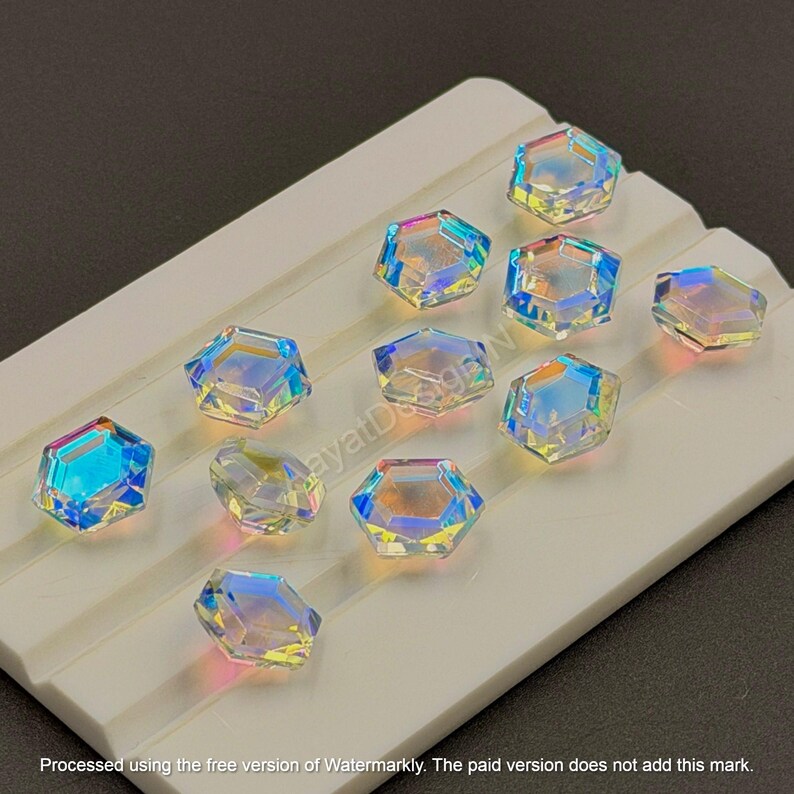 May include: Iridescent, hexagonal crystal beads arranged on a white surface. The crystals reflect a spectrum of colors, including blue, yellow, and pink. The beads are likely for jewelry making or craft projects.