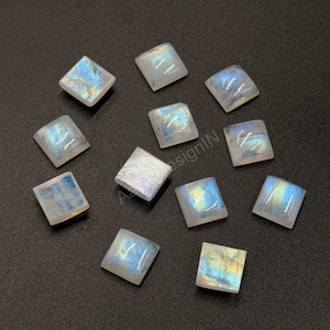 May include: A collection of eleven square-shaped moonstone cabochons arranged on a dark surface. Each gemstone displays a translucent, milky white appearance with iridescent blue and yellow flashes visible across their smooth, polished surfaces.