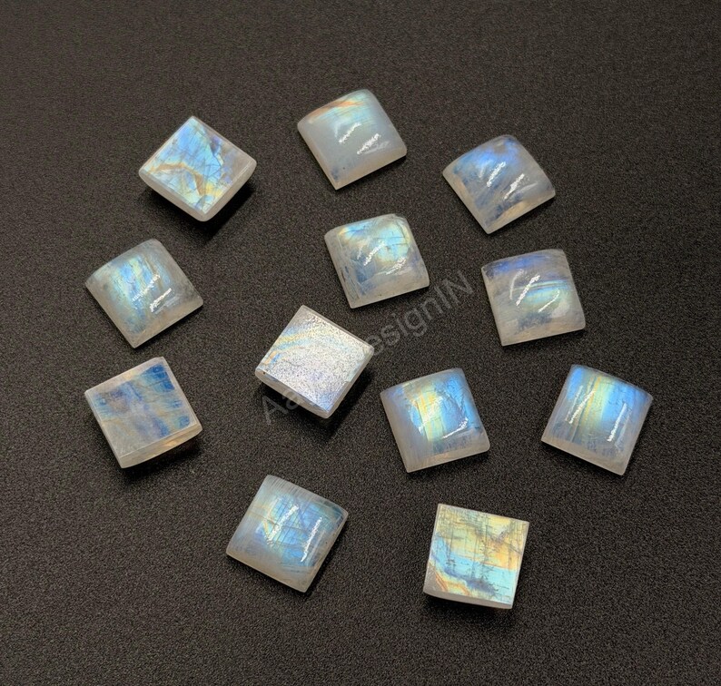May include: A collection of eleven square-shaped moonstone cabochons arranged on a dark surface. Each gemstone displays a translucent, milky white appearance with iridescent blue and yellow flashes visible across their smooth, polished surfaces.