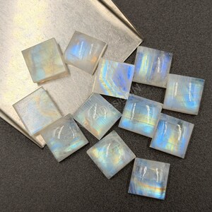 May include: A collection of eleven square-shaped moonstone cabochons featuring a translucent white base with iridescent blue and gold flashes. The stones are arranged in a scattered pattern, displaying smooth surfaces and sharp edges.