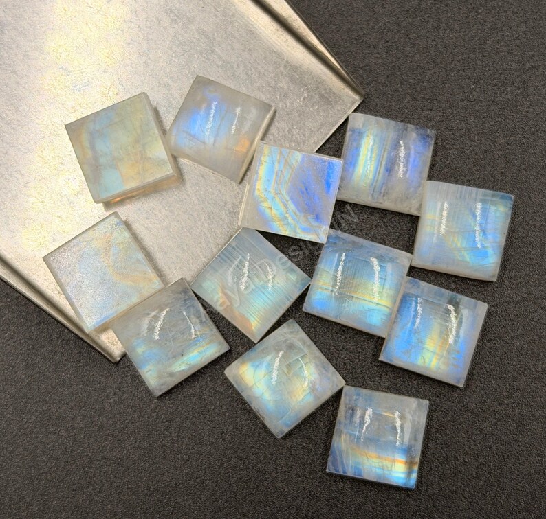 May include: A collection of eleven square-shaped moonstone cabochons featuring a translucent white base with iridescent blue and gold flashes. The stones are arranged in a scattered pattern, displaying smooth surfaces and sharp edges.