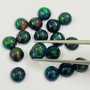 May include: A collection of round, black opal cabochons with green and red flashes of colour. The opals are loose and scattered on a white surface.