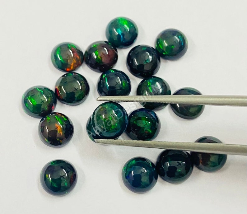 May include: A collection of round, black opal cabochons with green and red flashes of colour. The opals are loose and scattered on a white surface.