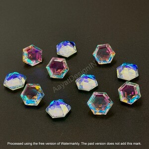 May include: Iridescent, faceted, hexagonal rhinestones scattered on a dark surface. The gems reflect a spectrum of colors, including pink, blue, and yellow. These decorative embellishments are suitable for various craft projects.
