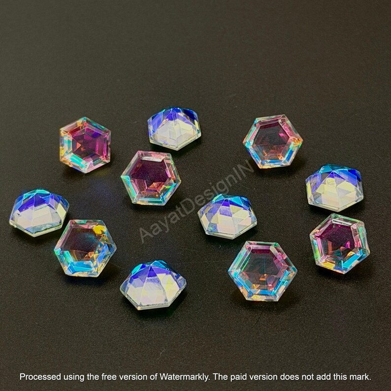 May include: Iridescent, faceted, hexagonal rhinestones scattered on a dark surface. The gems reflect a spectrum of colors, including pink, blue, and yellow. These decorative embellishments are suitable for various craft projects.