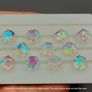 May include: Iridescent, hexagonal-shaped gemstones arranged on a white surface. The gems reflect a spectrum of colors, including blue, pink, and yellow. The gems are likely for jewelry making or crafting.