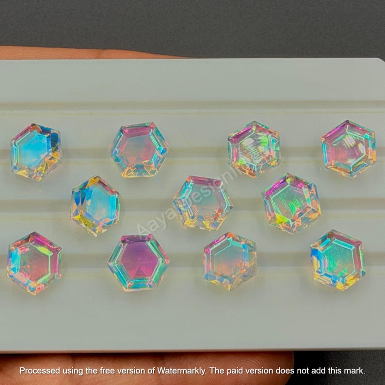 May include: Iridescent, hexagonal-shaped gemstones arranged on a white surface. The gems reflect a spectrum of colors, including blue, pink, and yellow. The gems are likely for jewelry making or crafting.
