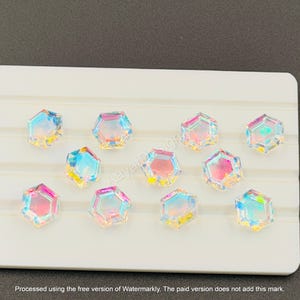 May include: Iridescent, hexagon-shaped crystal embellishments arranged on a white surface. The crystals reflect a spectrum of colors, including pink, blue, and yellow. The image includes the text "AayatDesign" and a watermark.