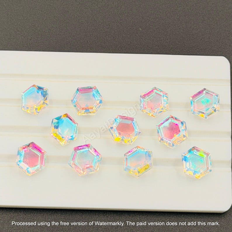May include: Iridescent, hexagon-shaped crystal embellishments arranged on a white surface. The crystals reflect a spectrum of colors, including pink, blue, and yellow. The image includes the text "AayatDesign" and a watermark.