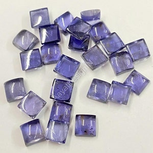 Peut inclure: Une collection de 18 petites pierres précieuses carrées, facettées et violettes. Les pierres précieuses sont disposées de manière aléatoire sur une surface blanche.