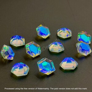 May include: A collection of iridescent, faceted crystal rhinestones. Each gem has a hexagonal shape with a flat back and a multi-colored, reflective surface. The crystals are scattered on a dark surface. Text at the bottom reads: Processed using the free version of Watermarkly.