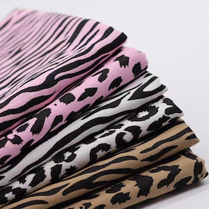 May include: A stack of folded fabric pieces featuring various animal print patterns. The collection includes pink, white, and tan base colours, each decorated with black zebra stripes or leopard spots, showcasing a textured weave throughout the fabric.