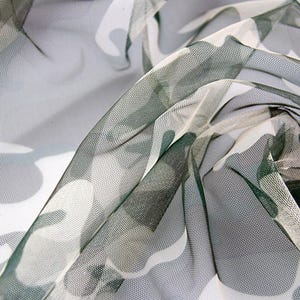 May include: A sheer, lightweight mesh fabric featuring a camouflage pattern in shades of dark green, light gray, and white. The material has a fine, semi-transparent grid texture with soft folds and drapes that create depth and visual movement.