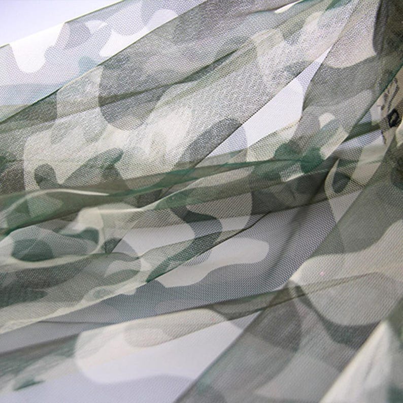 May include: A sheer, lightweight fabric featuring a camouflage pattern in shades of olive green, dark green, and white. The material has a fine, semi-transparent mesh texture with visible folds and drapes that create depth and layered shadows.