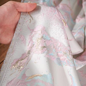 Puede incluir: Una pieza de tela con un tejido jacquard texturizado con un patr&oacute;n abstracto en tonos rosa suave, azul claro y oro met&aacute;lico sobre un fondo blanco. El material tiene un borde de orillo visible y una superficie tejida ligeramente elevada.