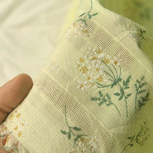 Op de afbeelding: Een lichtgele stof met een delicaat bloemmotief met witte en gele geborduurde bloesems en groene bedrukte stengels. Het textiel bevat horizontale banden met ajourwerk en een afwerking met een gerafelde rand.
