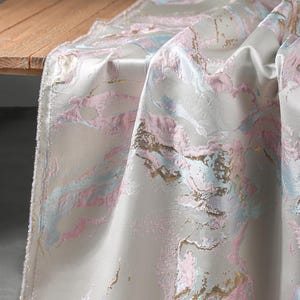 Puede incluir: Un camino de mesa de tela con un patr&oacute;n de m&aacute;rmol en tonos rosa suave, azul claro y oro met&aacute;lico sobre un fondo color crema. El material tiene un acabado liso y satinado con una apariencia texturizada y en relieve en las secciones de color.
