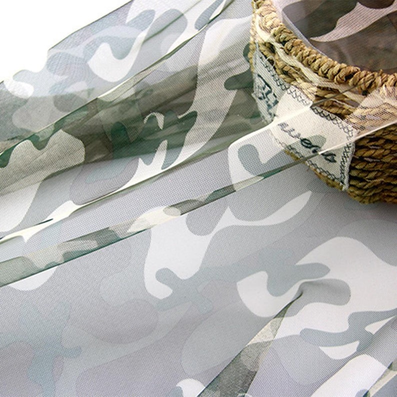 May include: A sheer, lightweight fabric featuring a camouflage pattern in shades of white, light gray, and dark olive green. The fabric is draped near a woven basket with a white fabric label that has the word "sweets" printed on it.