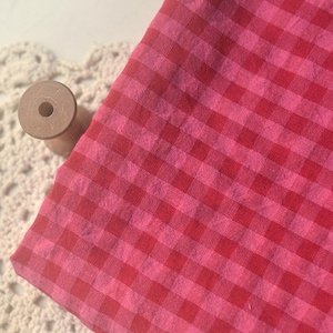 Op de afbeelding: Een stuk rood en roze geruit stof, opgevouwen. Een houten spoel ligt op een wit kanten kleedje. De stof heeft een klassiek geruit patroon in rood en roze.