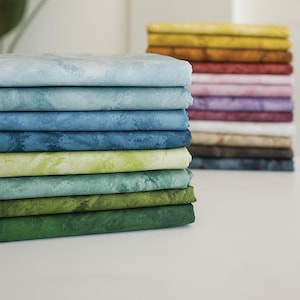 Puede incluir: Montones de tela doblada en varios colores, incluyendo azul claro, verde y azul oscuro. La tela tiene un aspecto veteado o texturizado. Otra pila de tela en tonos amarillos, rojos y morados es visible en el fondo.