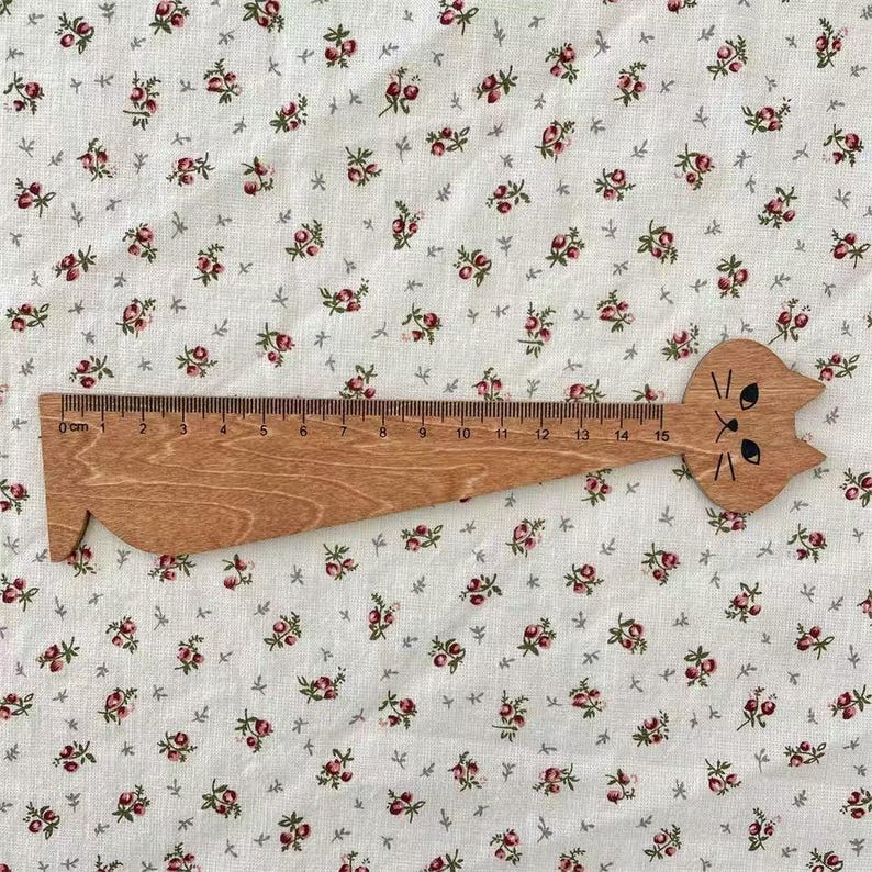 May include: A wooden ruler with a cat-shaped head. The ruler is brown and has black markings for centimetres, measuring up to 15 cm. The background is a white fabric with a floral pattern in pink and green.