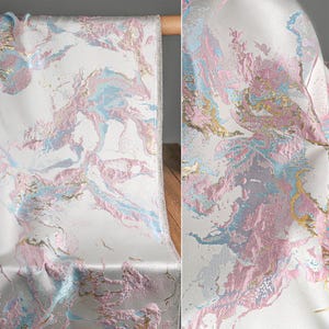 Puede incluir: Una tela con base blanca y un patr&oacute;n de m&aacute;rmol abstracto en tonos rosa pastel, azul claro y oro met&aacute;lico. El material tiene una apariencia tejida y texturizada con secciones elevadas que crean profundidad en toda la superficie.