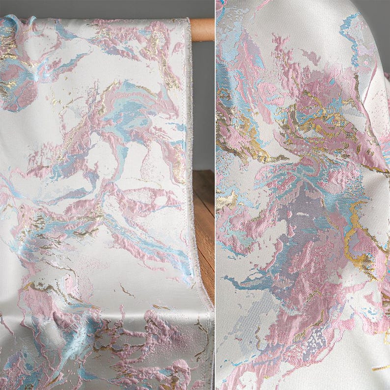 Puede incluir: Una tela con base blanca y un patr&oacute;n de m&aacute;rmol abstracto en tonos rosa pastel, azul claro y oro met&aacute;lico. El material tiene una apariencia tejida y texturizada con secciones elevadas que crean profundidad en toda la superficie.
