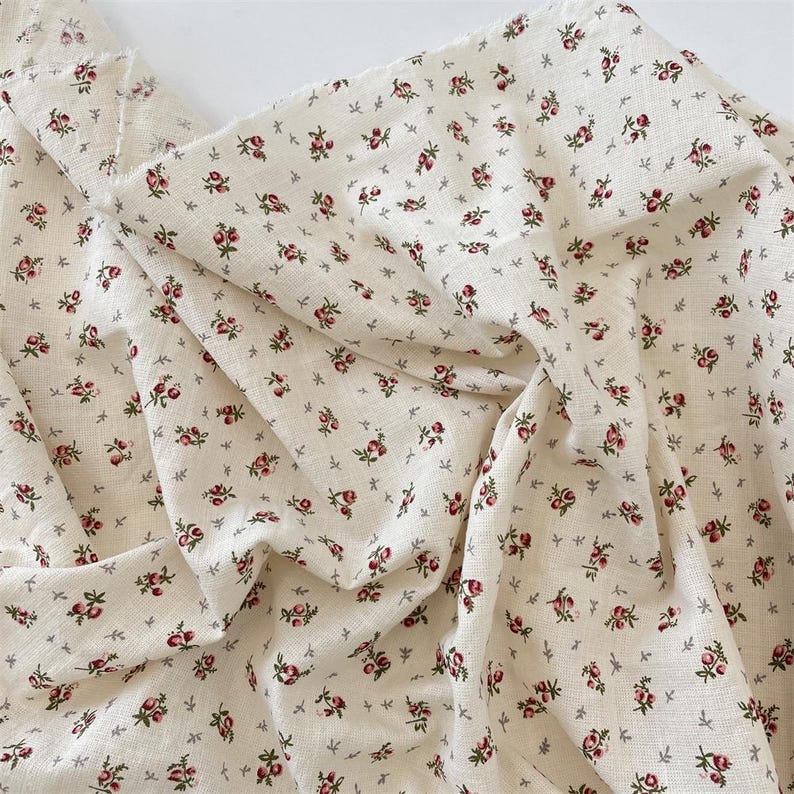 May include: Off-white fabric featuring a delicate floral pattern of tiny red roses and grey leaves. The material is creased, possibly linen or cotton. The design evokes a vintage or cottagecore style, suitable for various sewing projects or home decor.