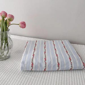 May include: Folded blue and white striped fabric with floral accents. A clear glass vase holds pink tulips, set against a white background. The fabric features vertical stripes and rows of small red flowers.