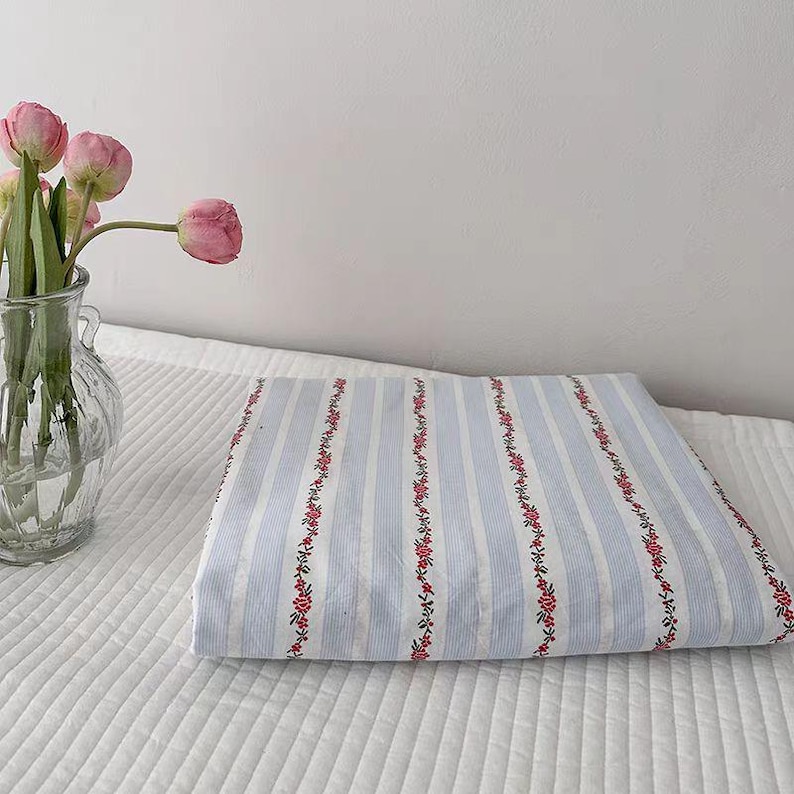 May include: Folded blue and white striped fabric with floral accents. A clear glass vase holds pink tulips, set against a white background. The fabric features vertical stripes and rows of small red flowers.
