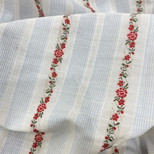 Pode incluir: Close-up de um tecido com um padrão listrado em azul claro e branco. Fileiras verticais de bordados florais vermelhos com folhas verdes adornam o tecido. O tecido está dobrado, revelando sua textura e design.